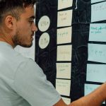 A man in a casual t-shirt organizes notes on a wall, focusing on a 'WORK OUT' sign.