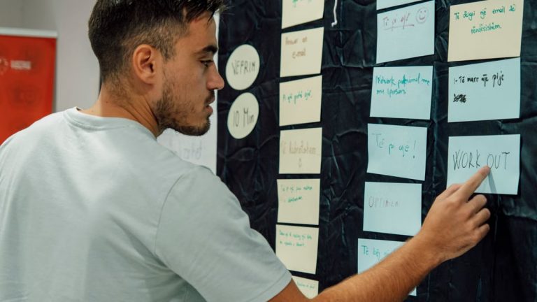 A man in a casual t-shirt organizes notes on a wall, focusing on a 'WORK OUT' sign.