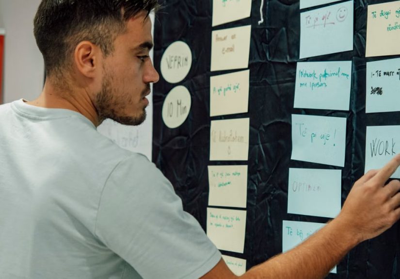 A man in a casual t-shirt organizes notes on a wall, focusing on a 'WORK OUT' sign.